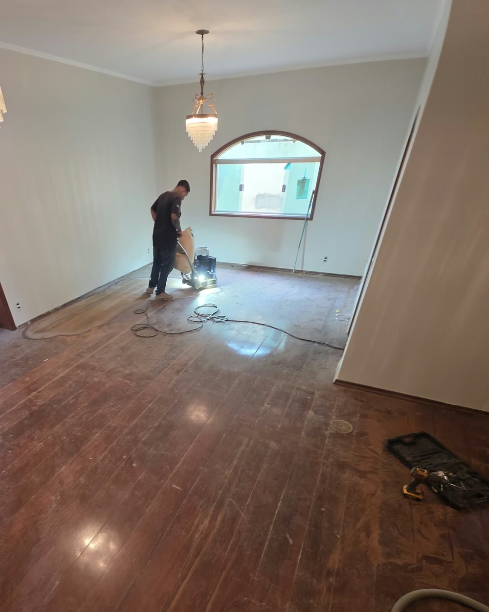 Leonardo Comino raspando o assoalho com a lixadeira ligada ao aspirador, em sala com lustre de cristal