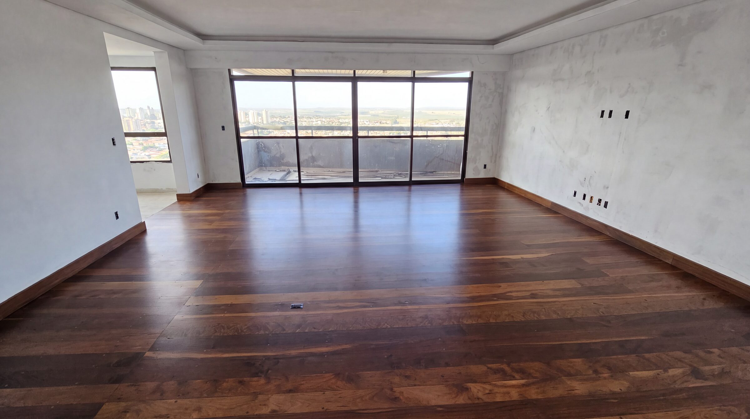 Sala vazia com assoalho maciço escuro instalado e parede de janelas com vista da cidade